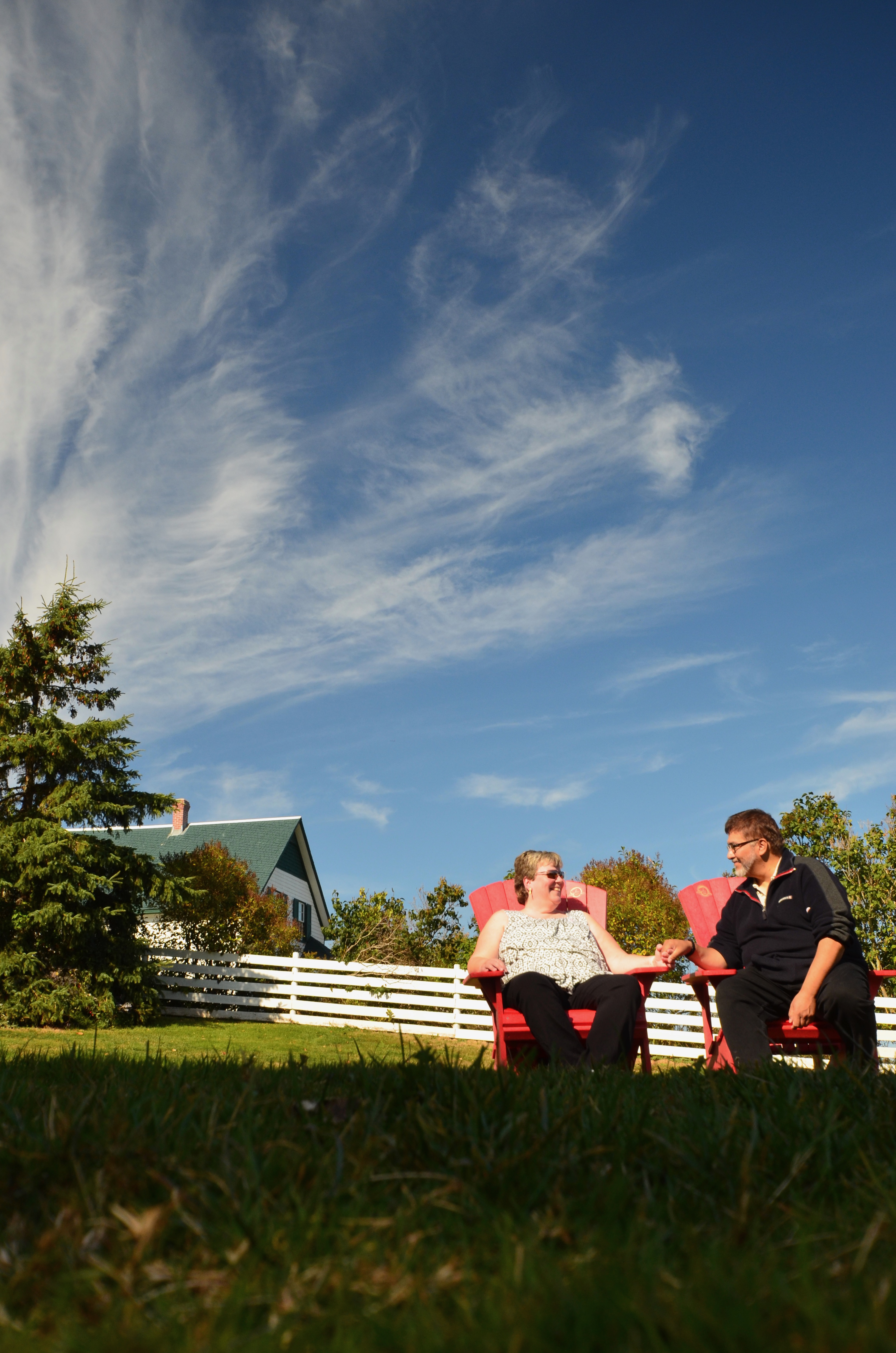 Couple sitting on red chairs in front of green house with white picket fence.